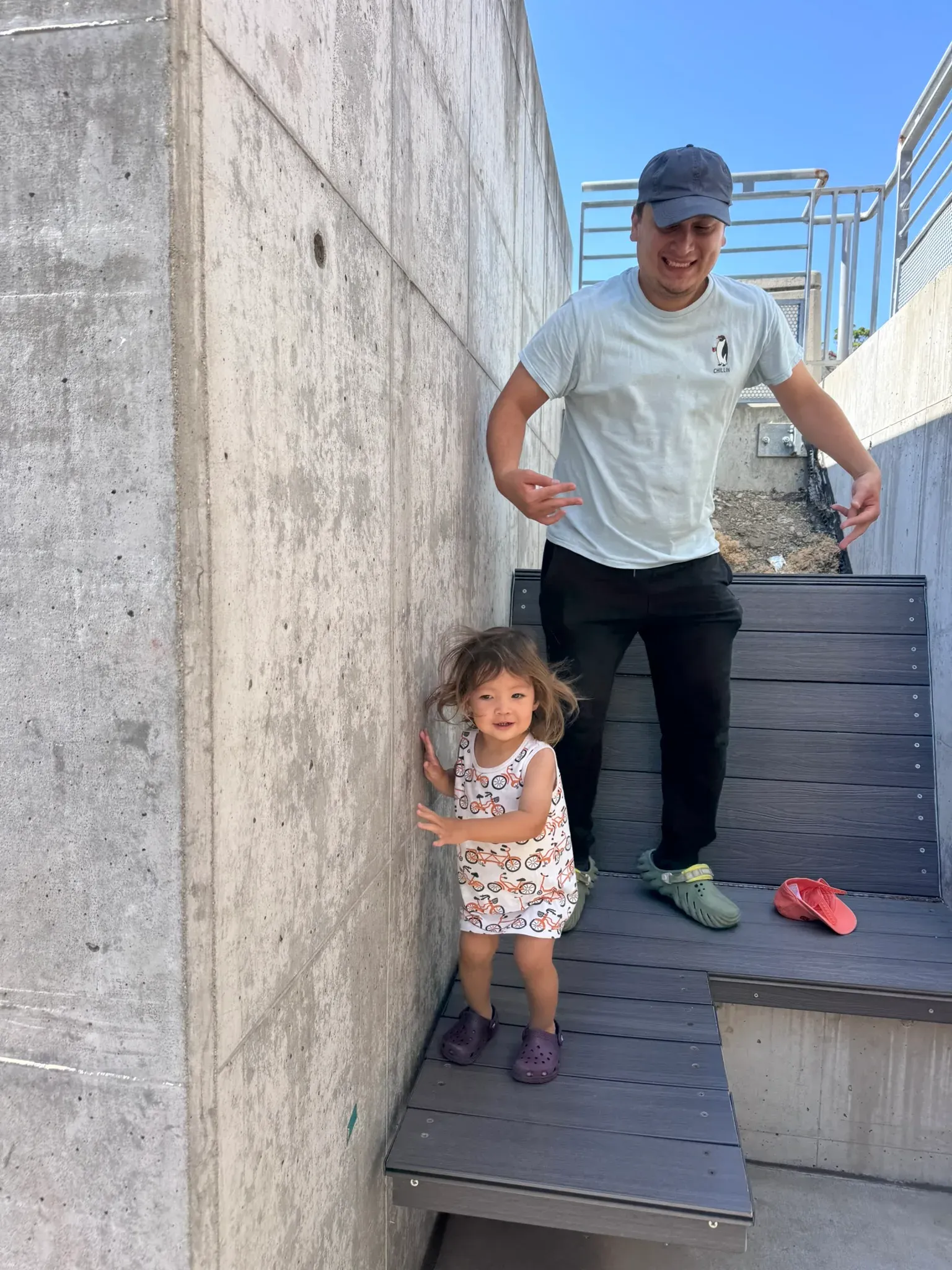 A man and a small child on the same bench on a bright day, the child standing on the deck mid-step with shoes left beside her