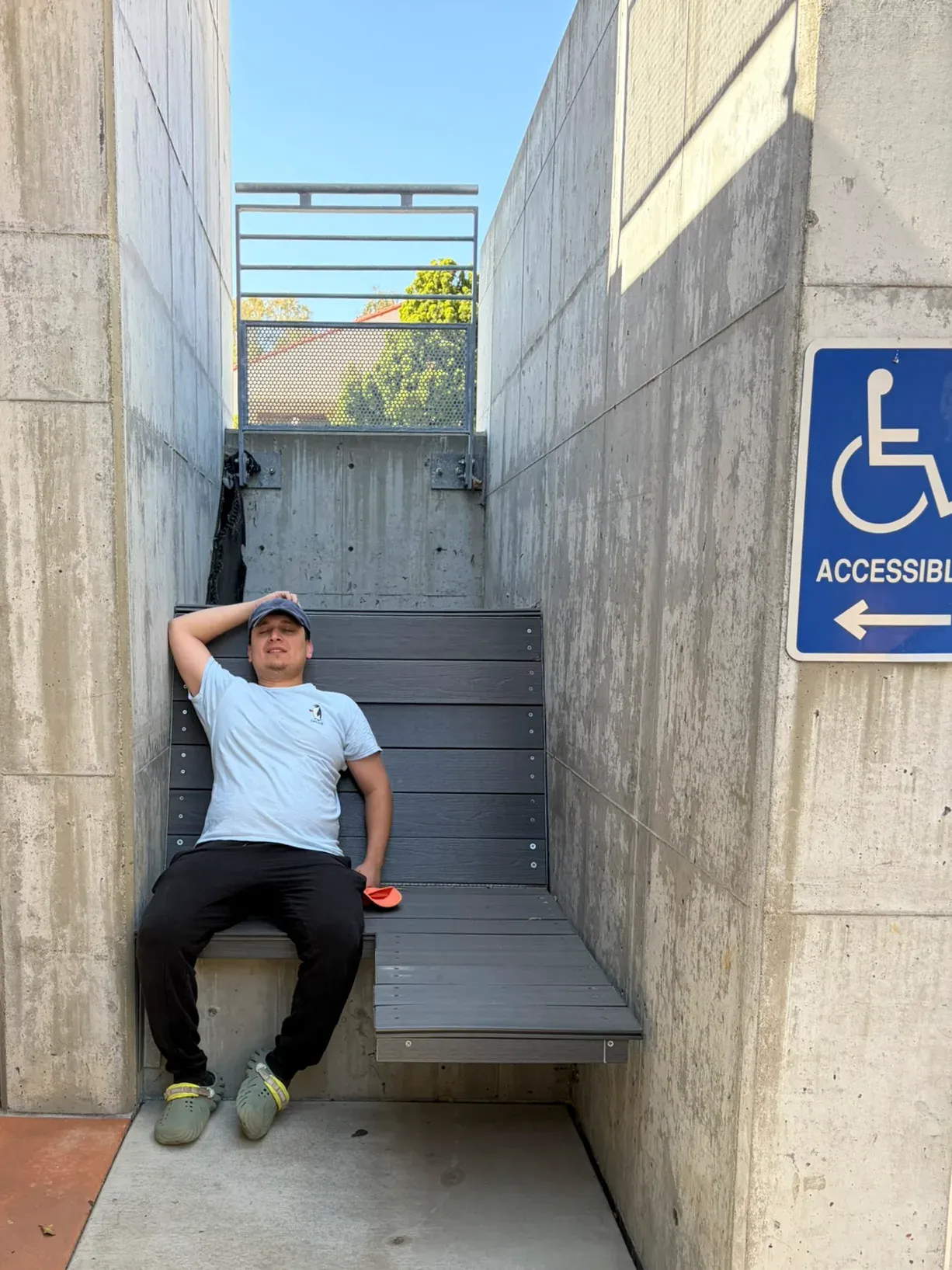 A man lying back along the finished bench in full sun, one hand on his cap and his shoes off beside him, the accessible route sign on the concrete pier to the right